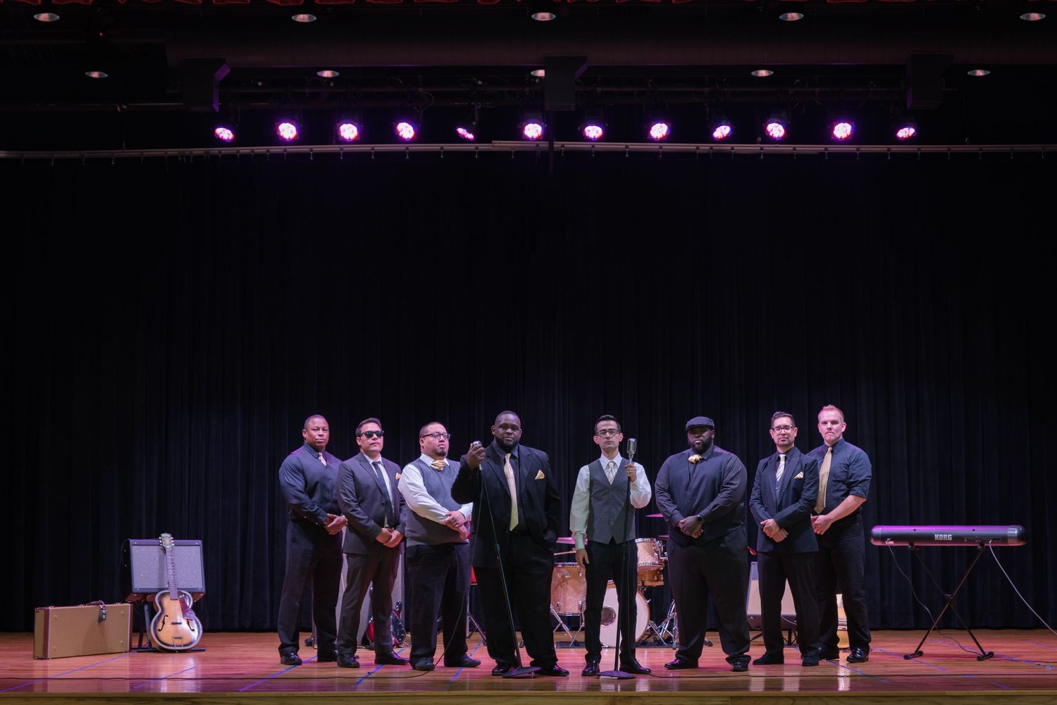 Performers in dark suits stand in formation on a dimly lit stage during a choral performance.