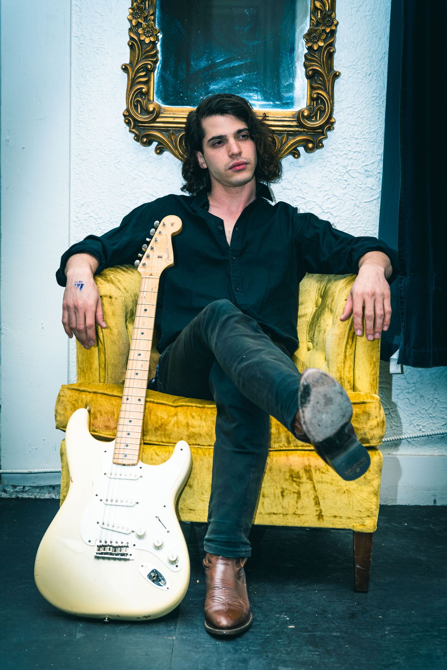 A musician lounges in a yellow vintage armchair with a white Fender Stratocaster electric guitar against a white wall.