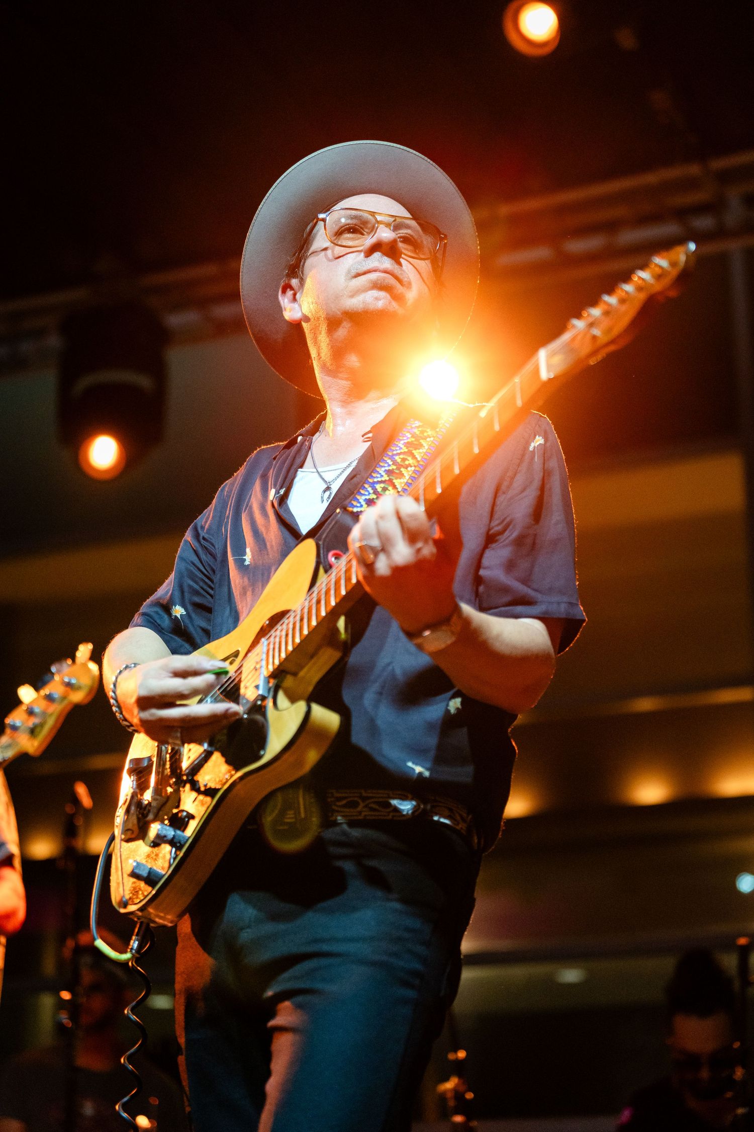 Musician in hat and blue shirt plays electric guitar on stage under dramatic orange lighting during performance.