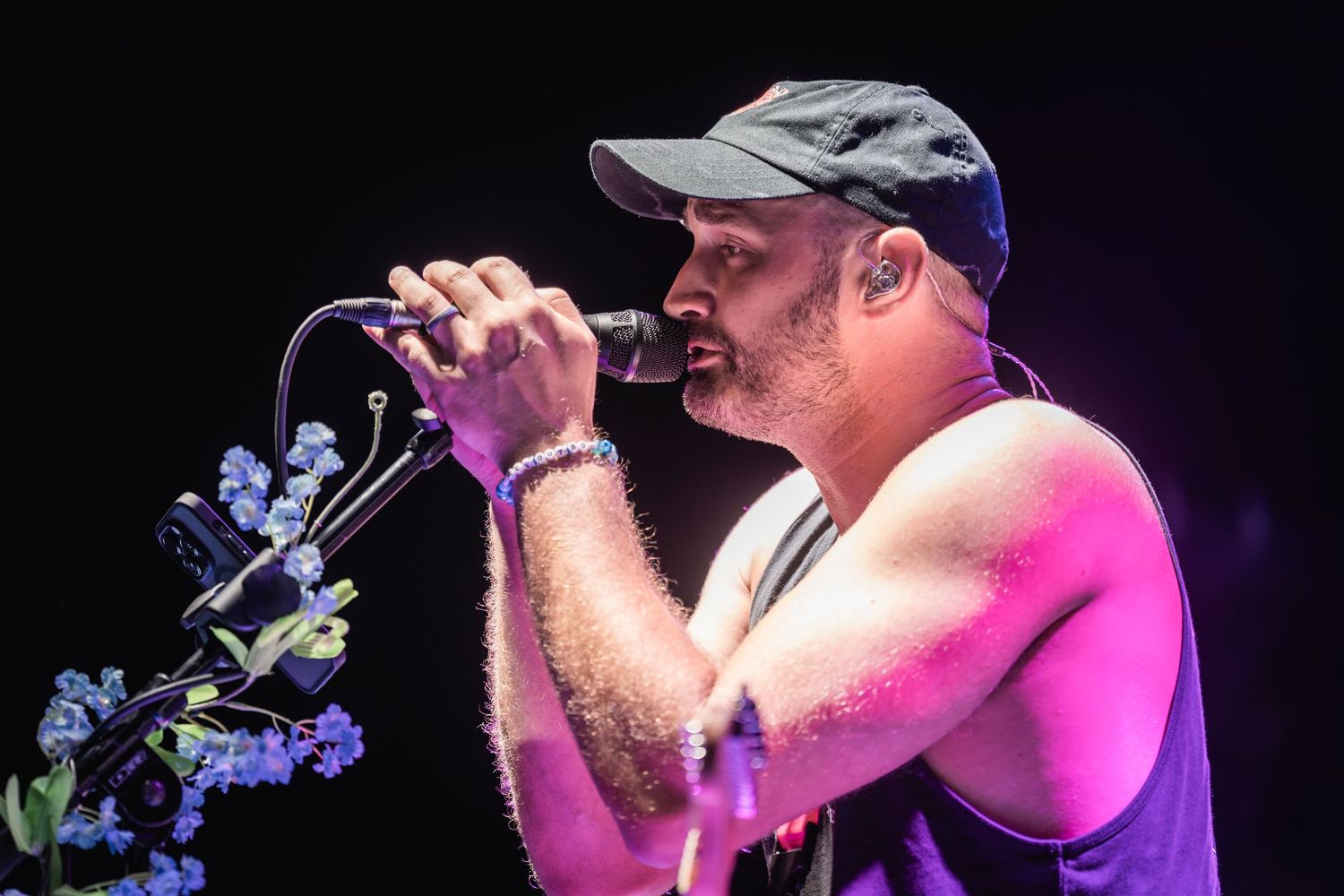 A performer in a baseball cap sings passionately on stage under colorful concert lighting during a live performance.