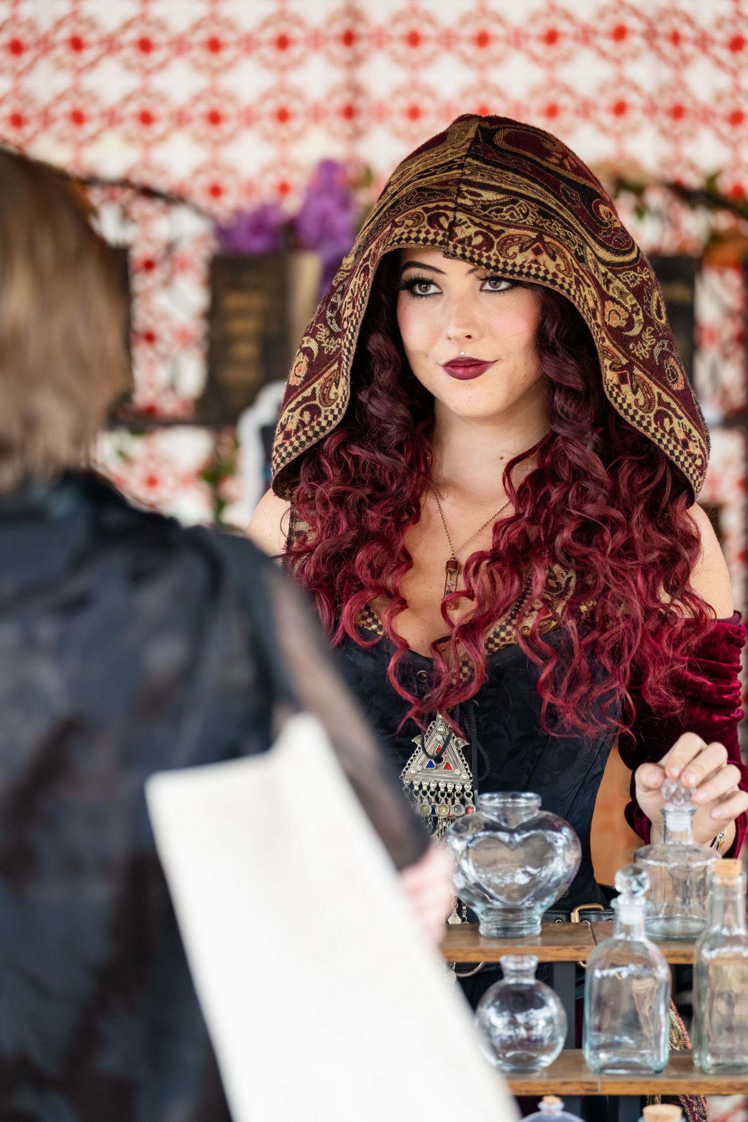 A person with vibrant red curls wearing an ornate hooded shawl sits at a table with glass jars in a decorated setting.