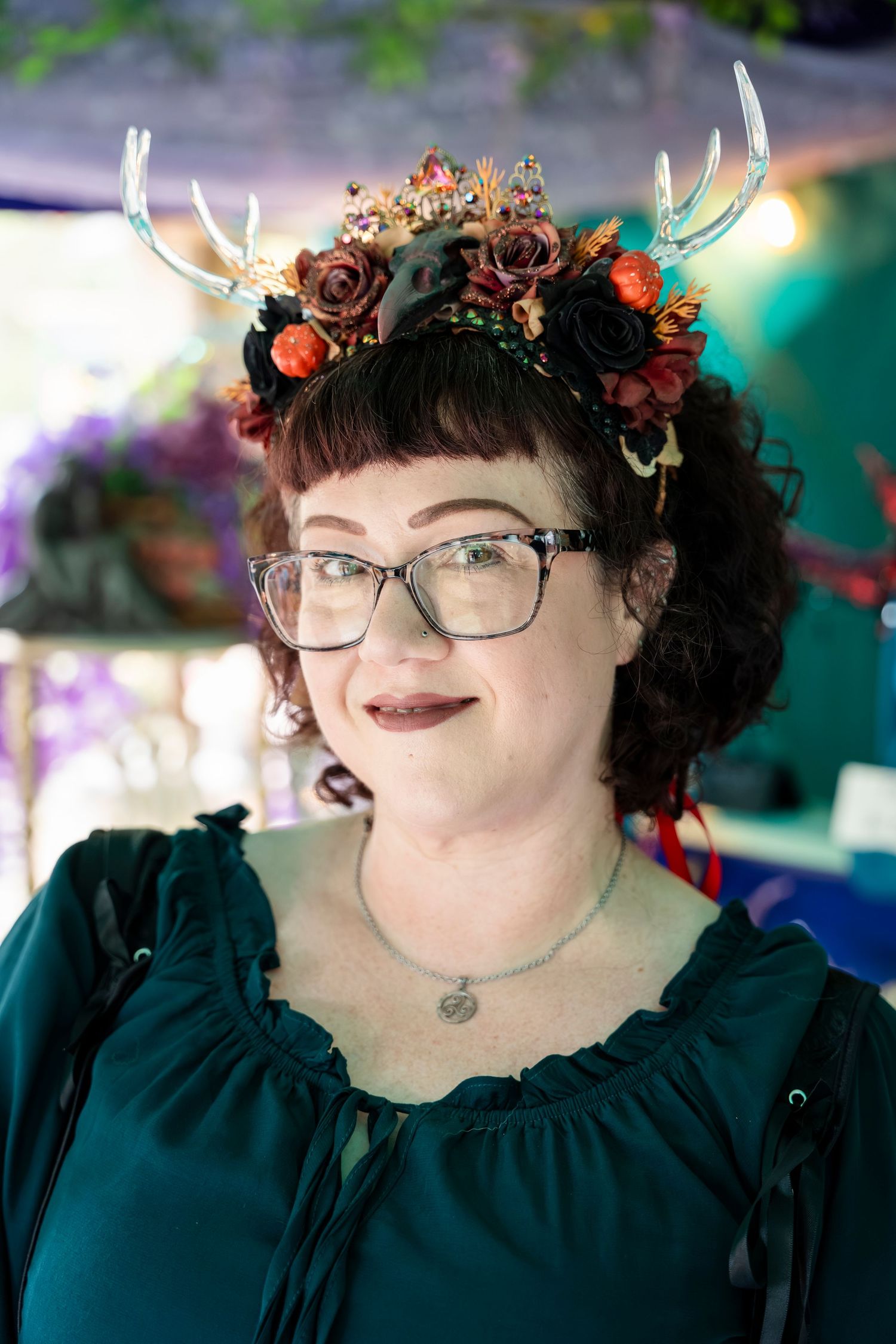 A whimsical floral crown with antlers adorns a person wearing glasses and a dark green top against a colorful background.