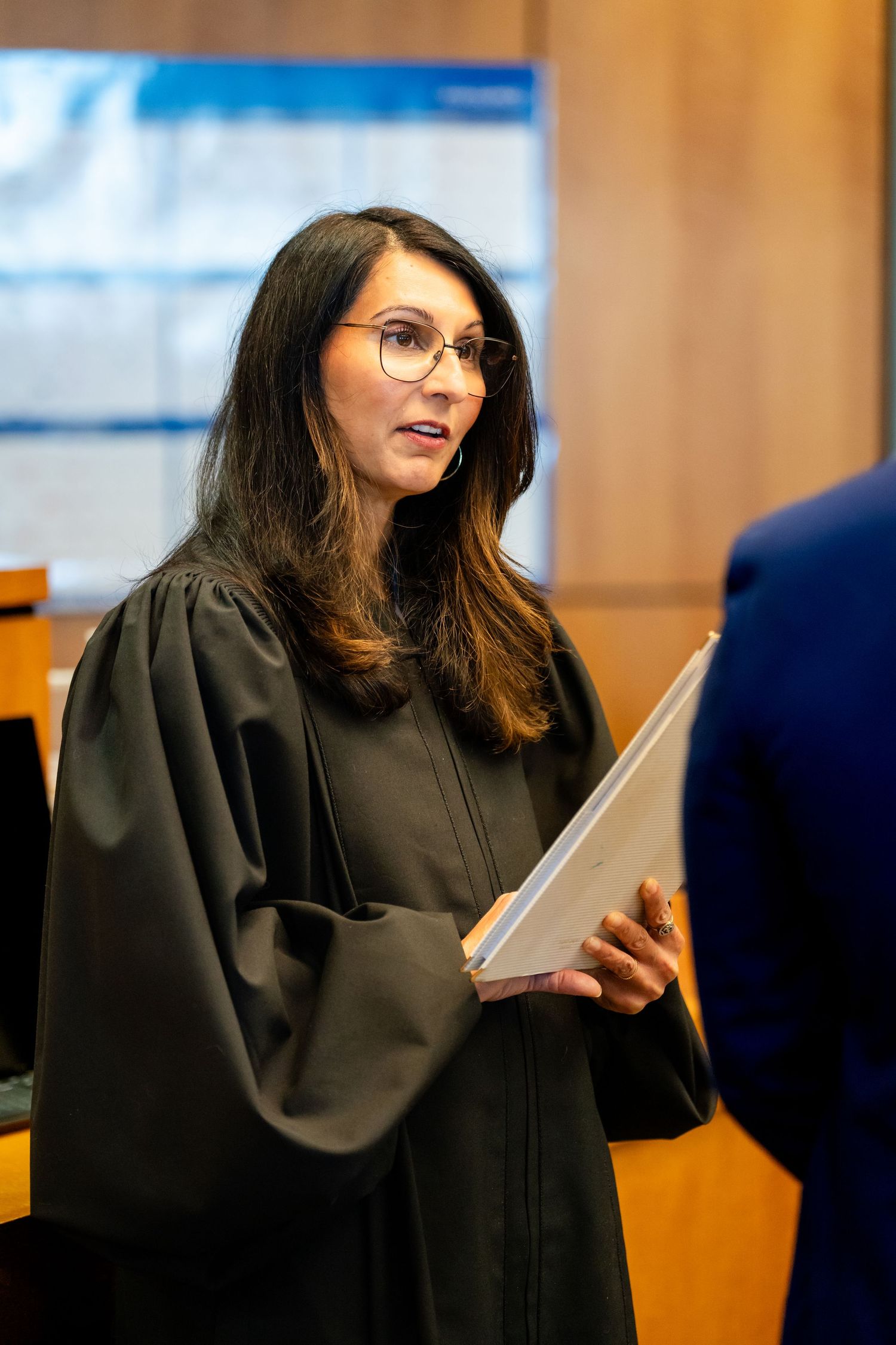 A person wearing a black judicial robe and glasses speaks while holding documents in a courtroom setting.