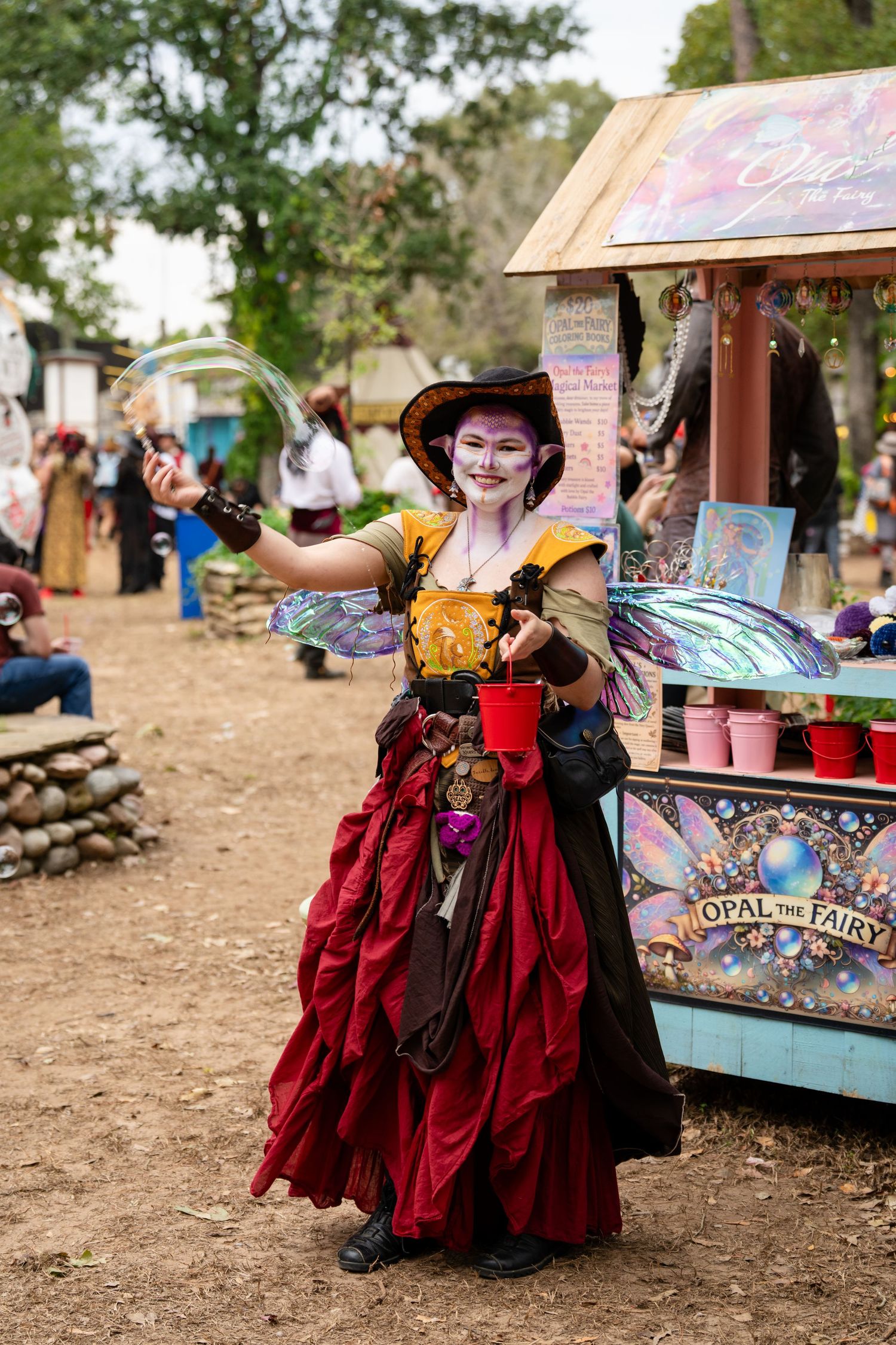 A performer in a burgundy Renaissance dress poses playfully near vendor stalls at a medieval festival.