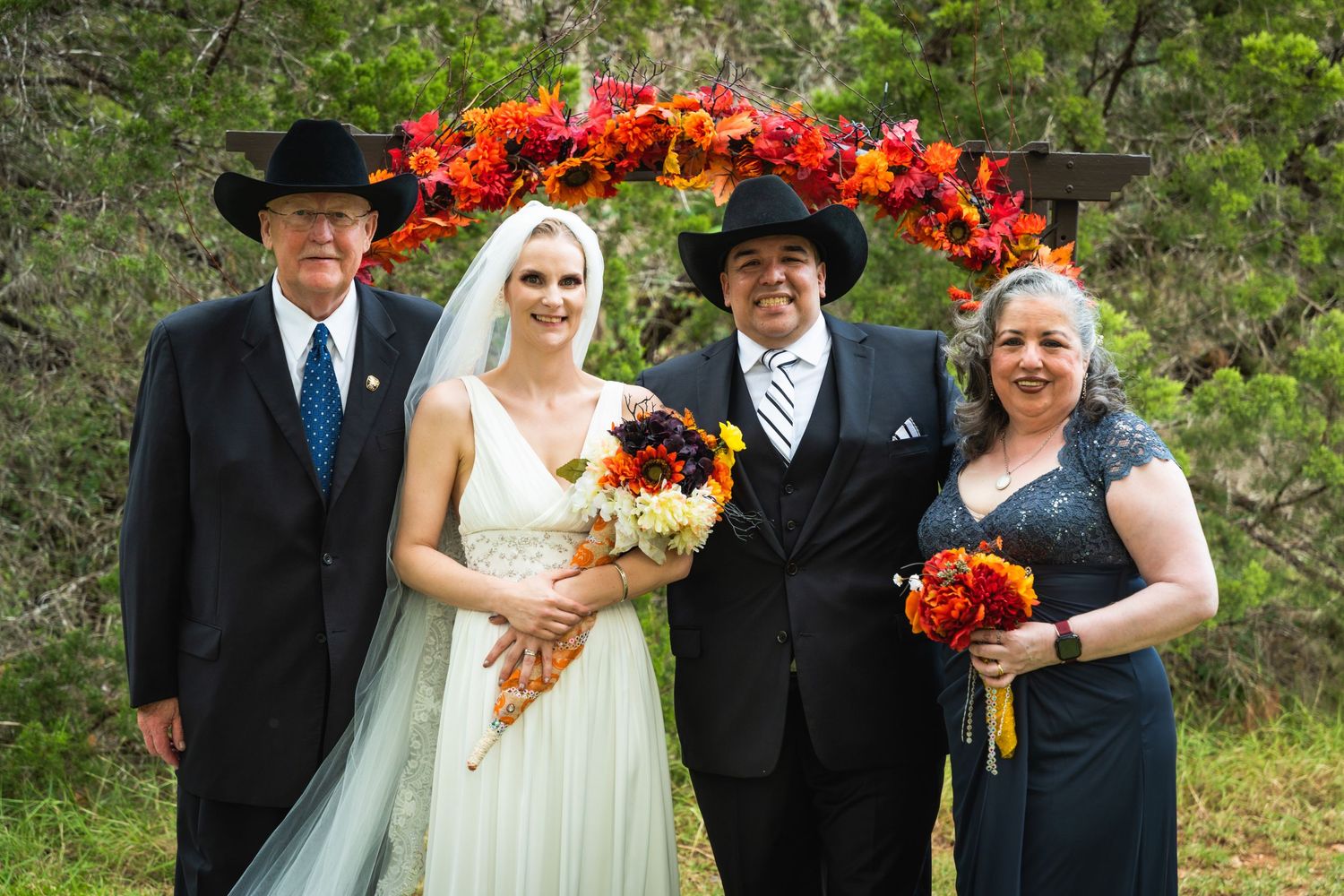 Wedding party poses beneath a bright red floral arch with autumn-themed bouquets.