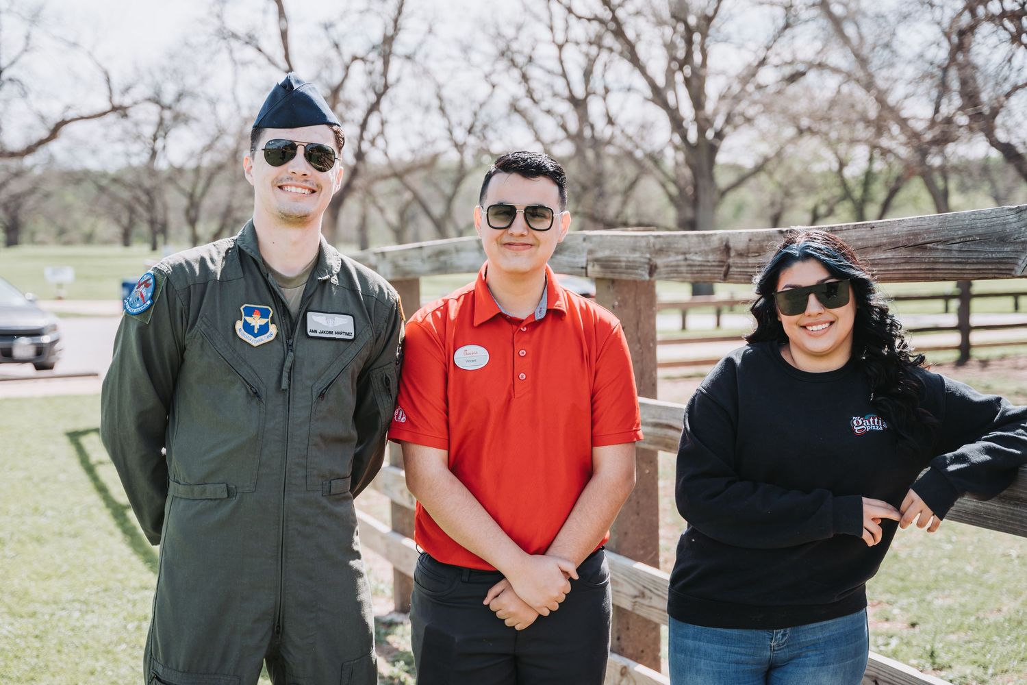 Group of three people standing outdoors near a rustic fence wearing casual attire and sunglasses.