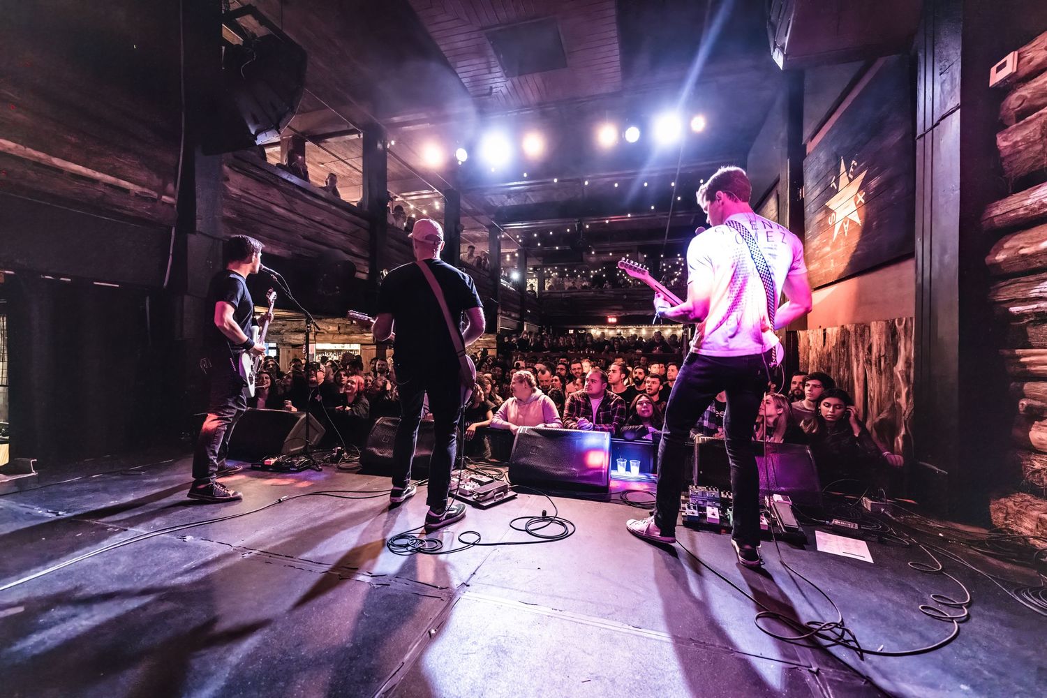 Band performs live on wooden-walled stage with blue stage lighting and crowd visible.