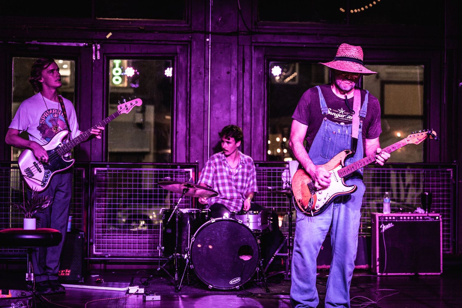 Three-piece band performs on outdoor stage with purple lighting effects.