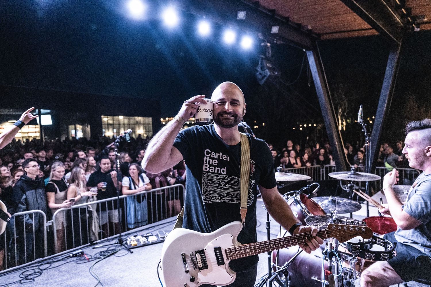 Musician performs with guitar on outdoor stage with bright lighting and crowd barriers.