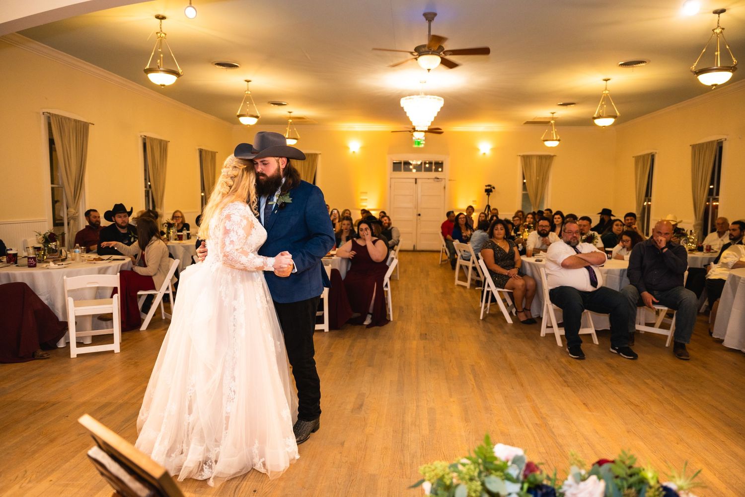 Couple sharing their first dance in an elegant ballroom with guests watching from tables.
