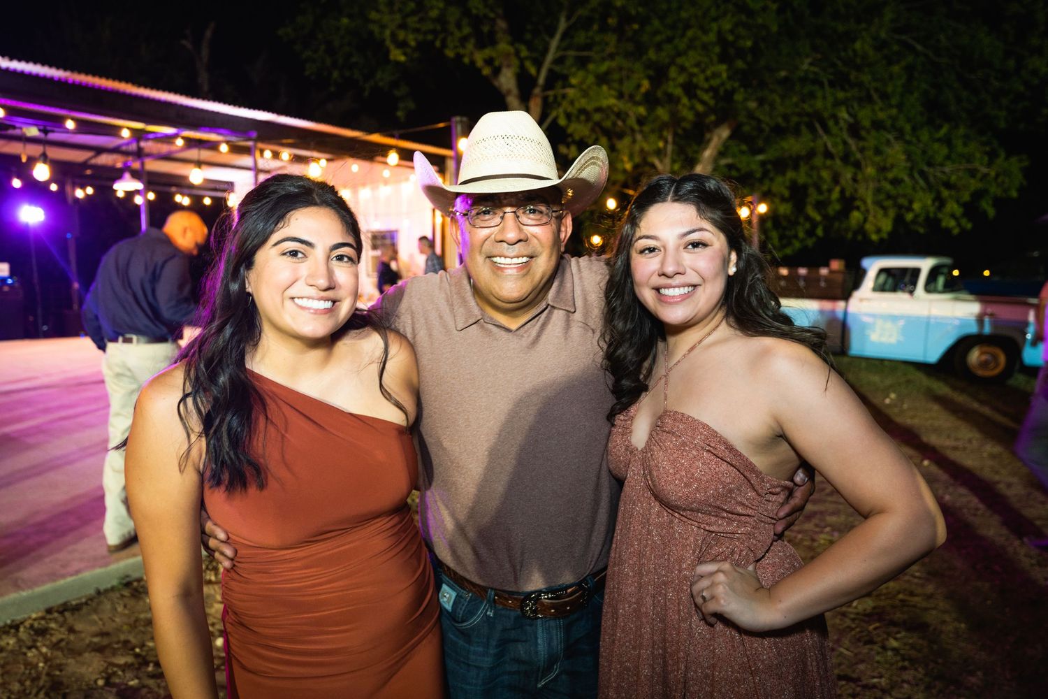 Three people posing together at a western-themed outdoor party with string lights overhead.