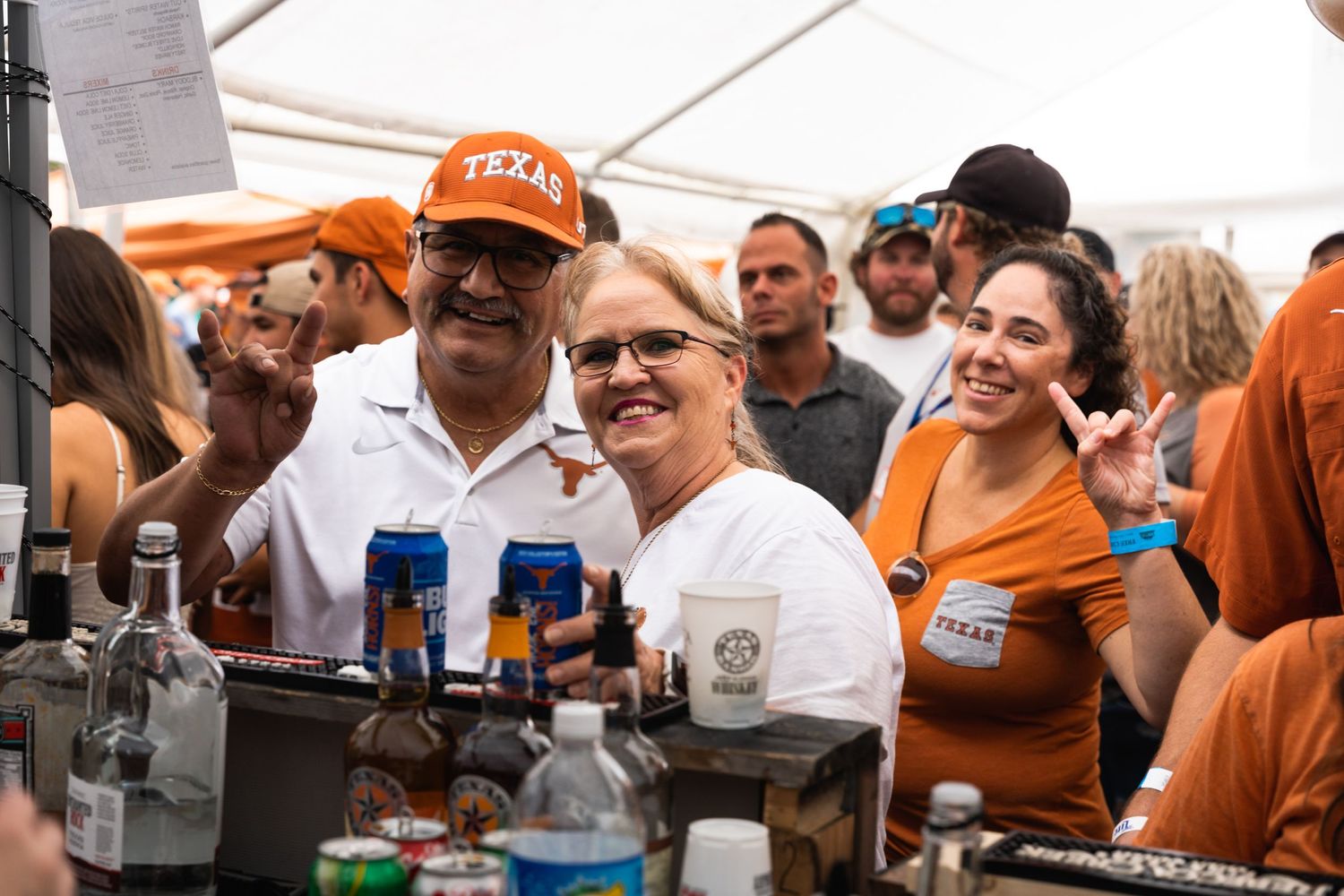 People enjoying drinks and socializing under a white tent at an outdoor event with orange and blue decorations.