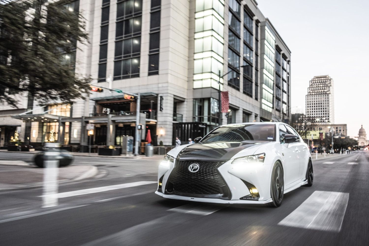 Modified white Lexus sedan parked on urban street corner with high-rise buildings.