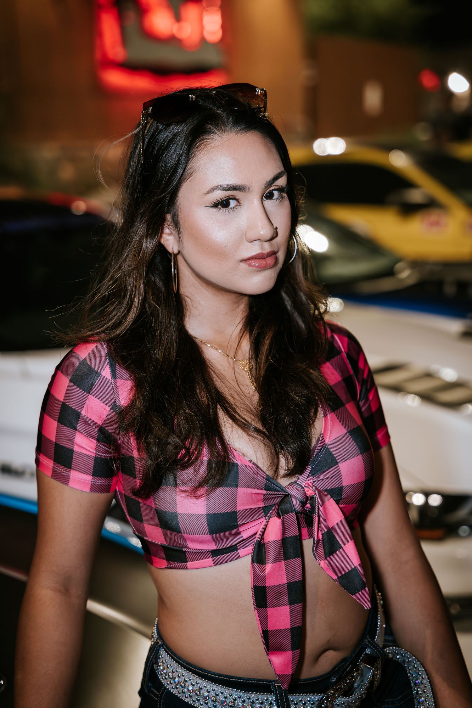 Model in pink buffalo check crop top poses at nighttime car show.
