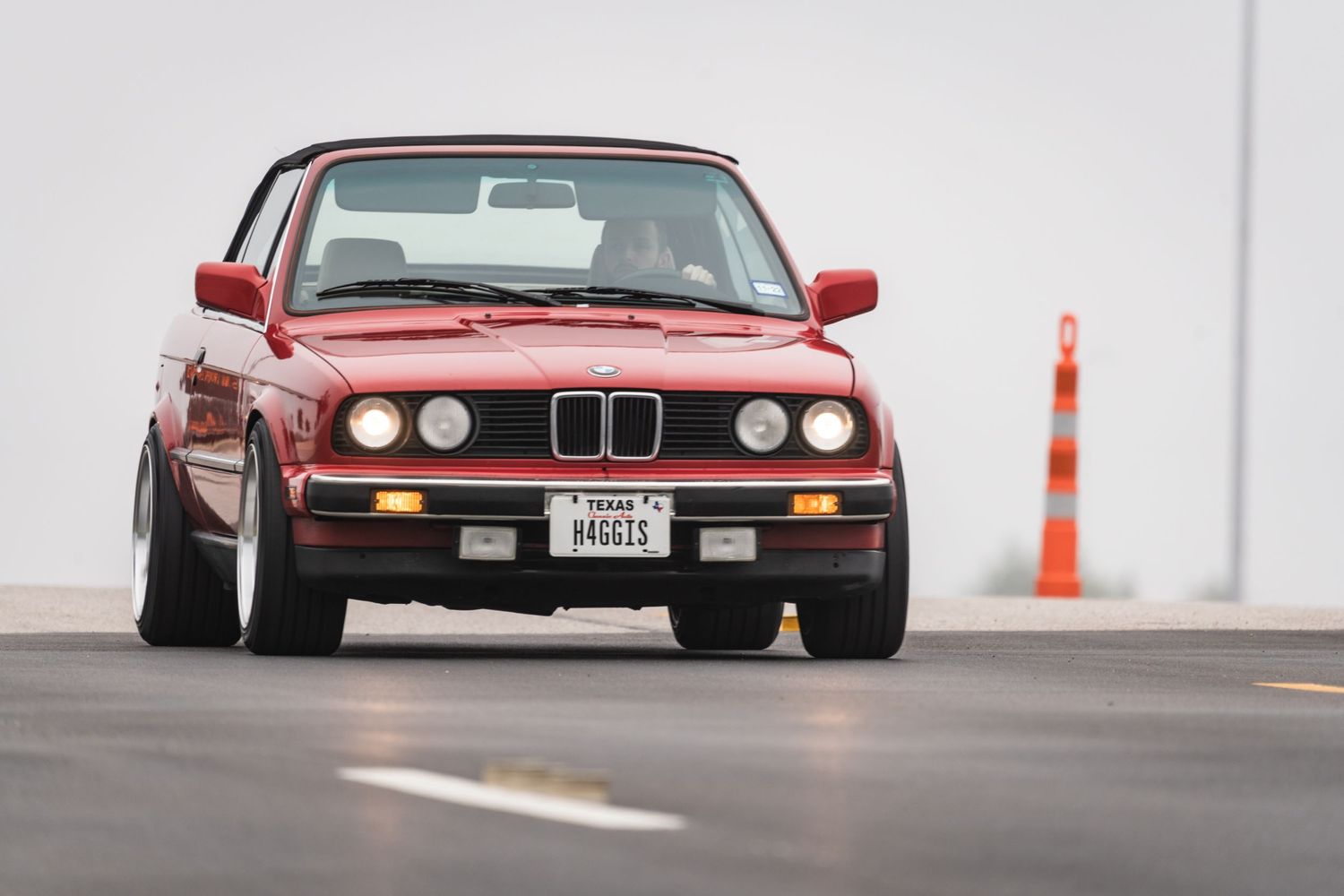 Red BMW E30 sports car taking a corner on a race track with orange cones.