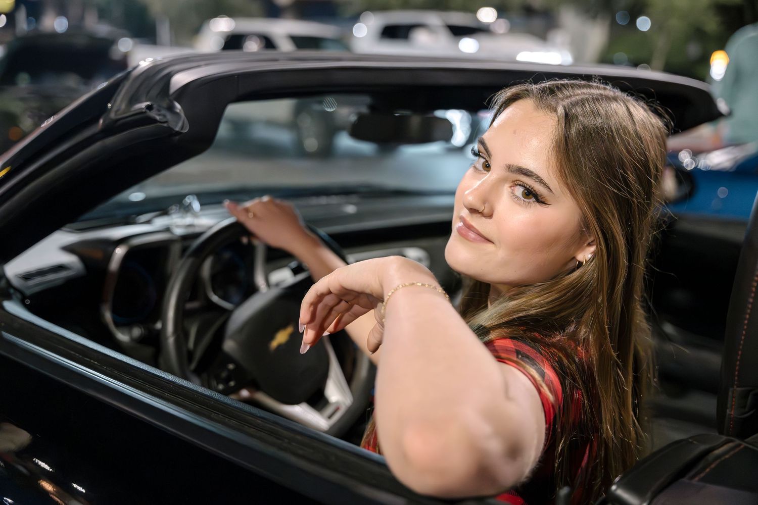 A person poses behind the wheel of a black convertible car at twilight.