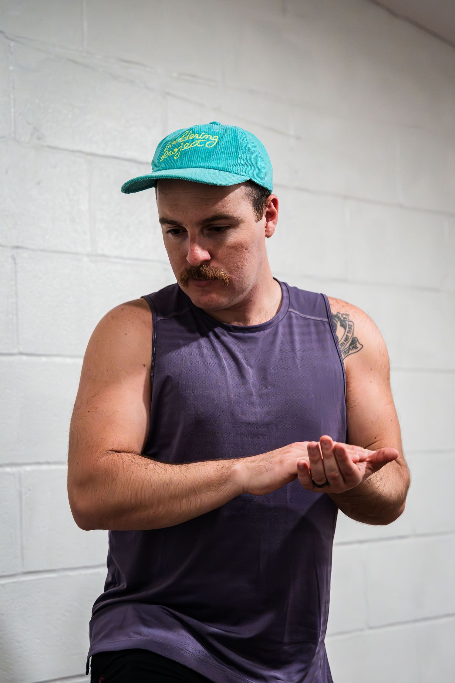 A musician in a purple tank top and teal cap stands in front of a white wall.