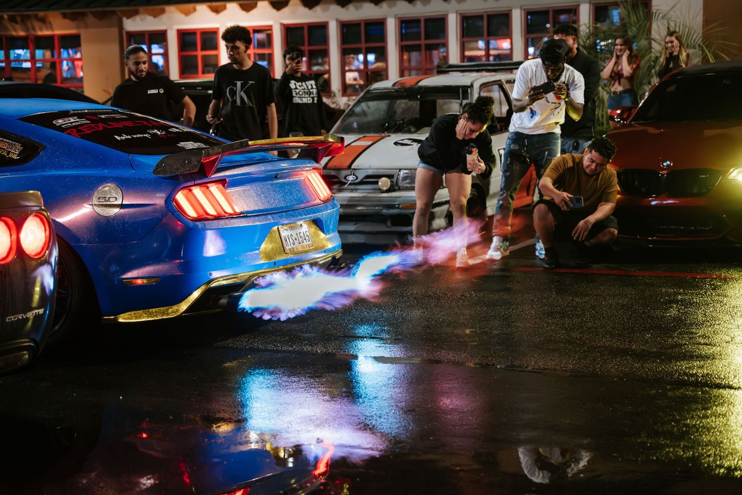 Blue sports car backfiring with flames reflecting in wet pavement at a nighttime car meet.