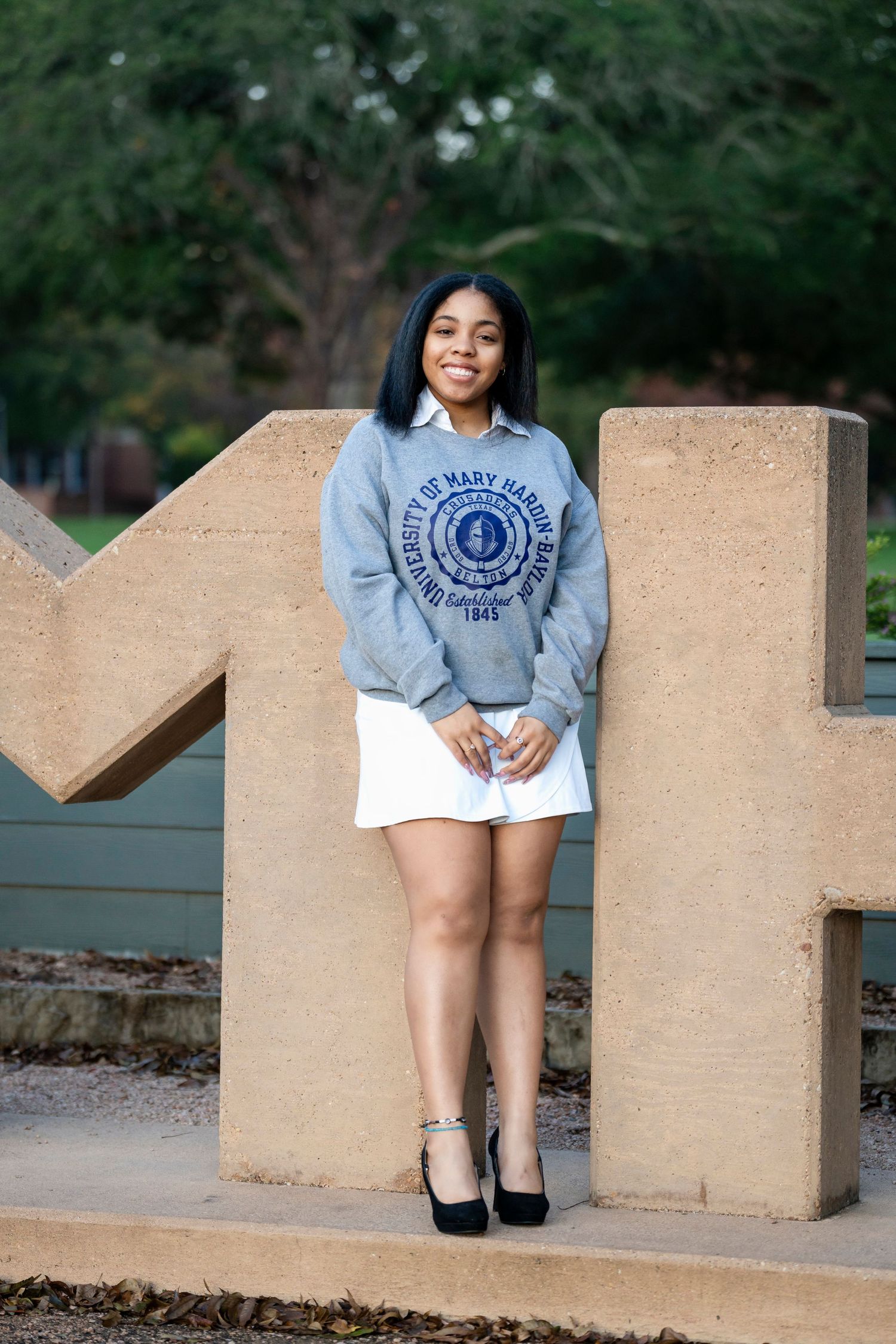 A student in university sweatshirt poses by concrete letter M on campus.
