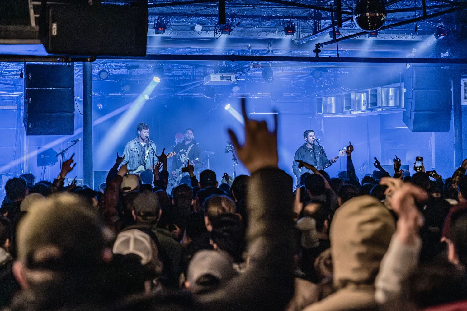 Enthusiastic crowd at concert venue with raised hands watching band perform under blue lights.