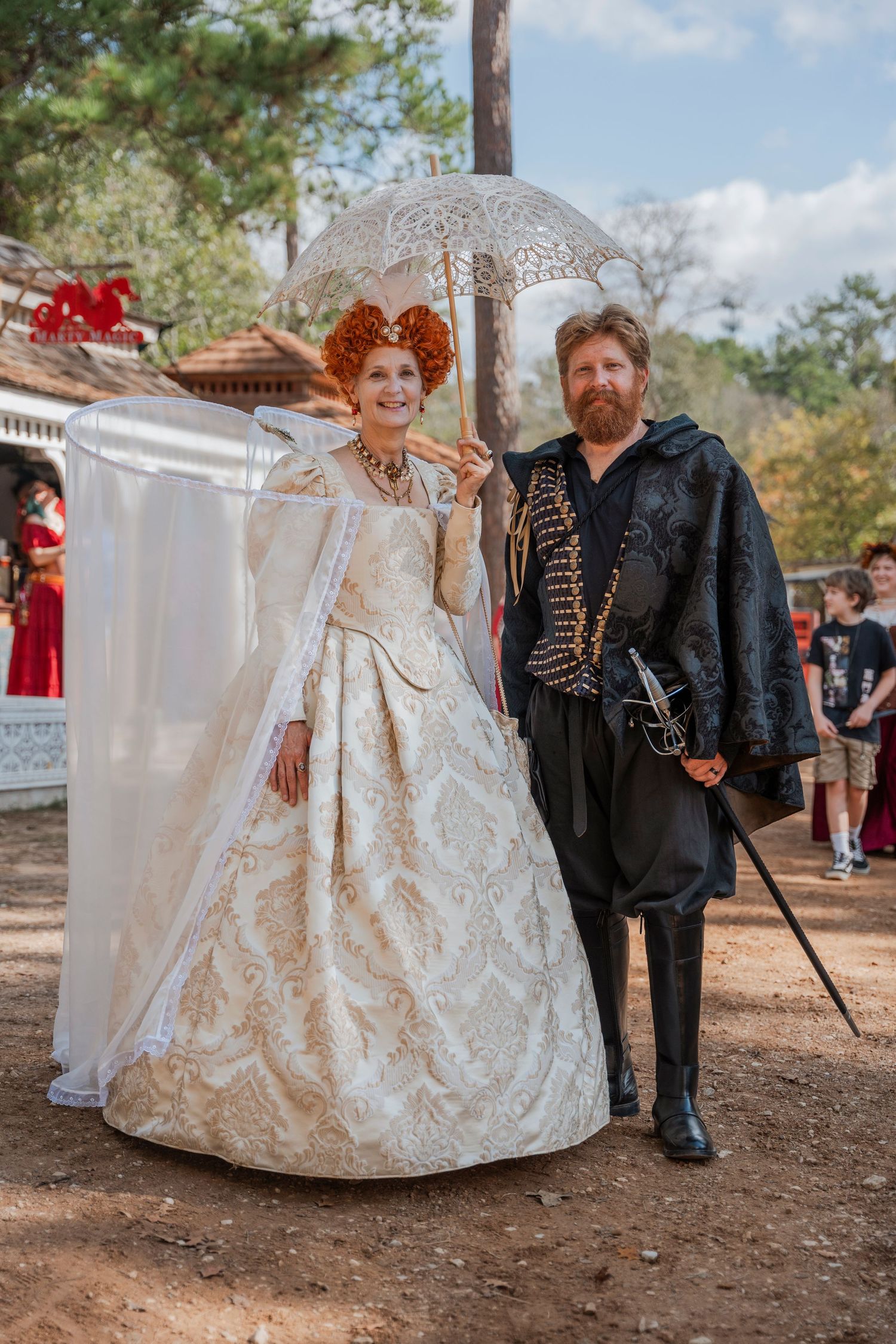 A couple in elegant renaissance period costumes poses with a white parasol at an outdoor fair.