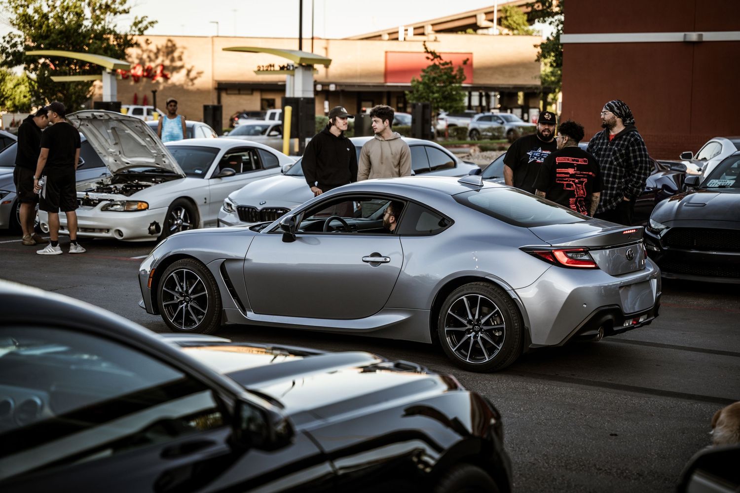 Silver sports car parked in a crowded lot at sunset with other vehicles and figures visible.