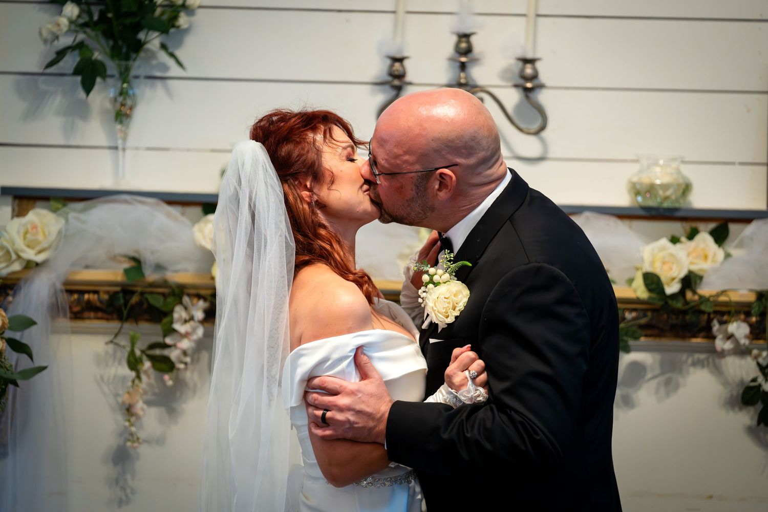A wedding couple shares their first kiss in a decorated room with white walls and floral arrangements.