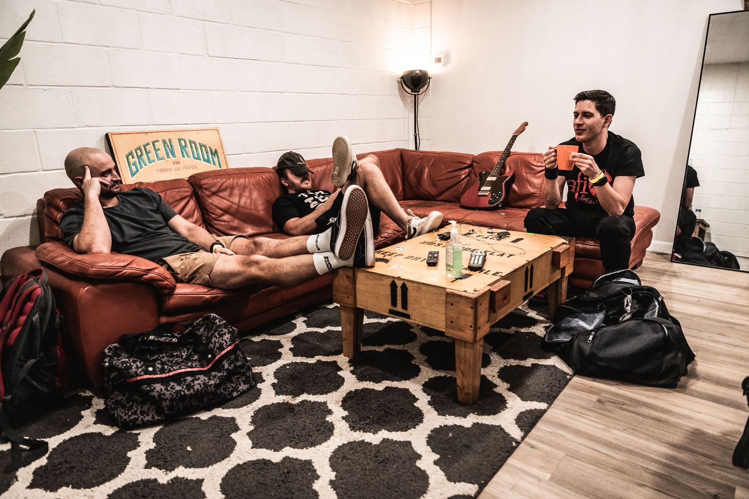 Musicians relax on a brown leather couch in a living room setting.