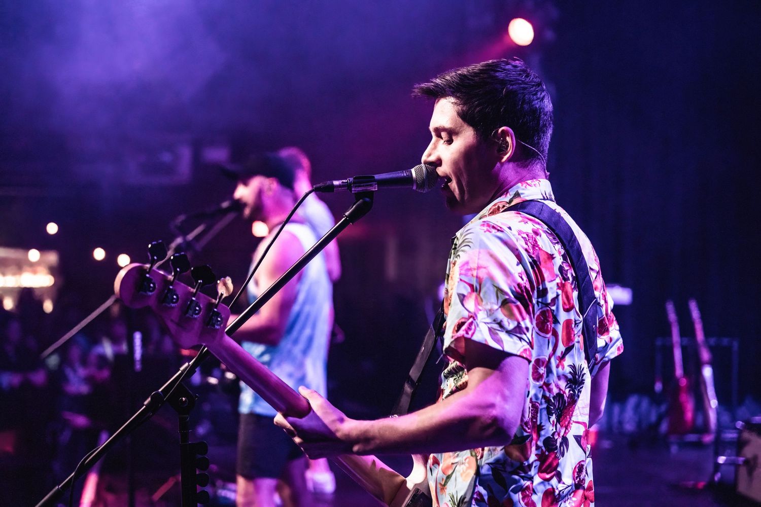 Musician performs on stage wearing a floral shirt under purple concert lighting.