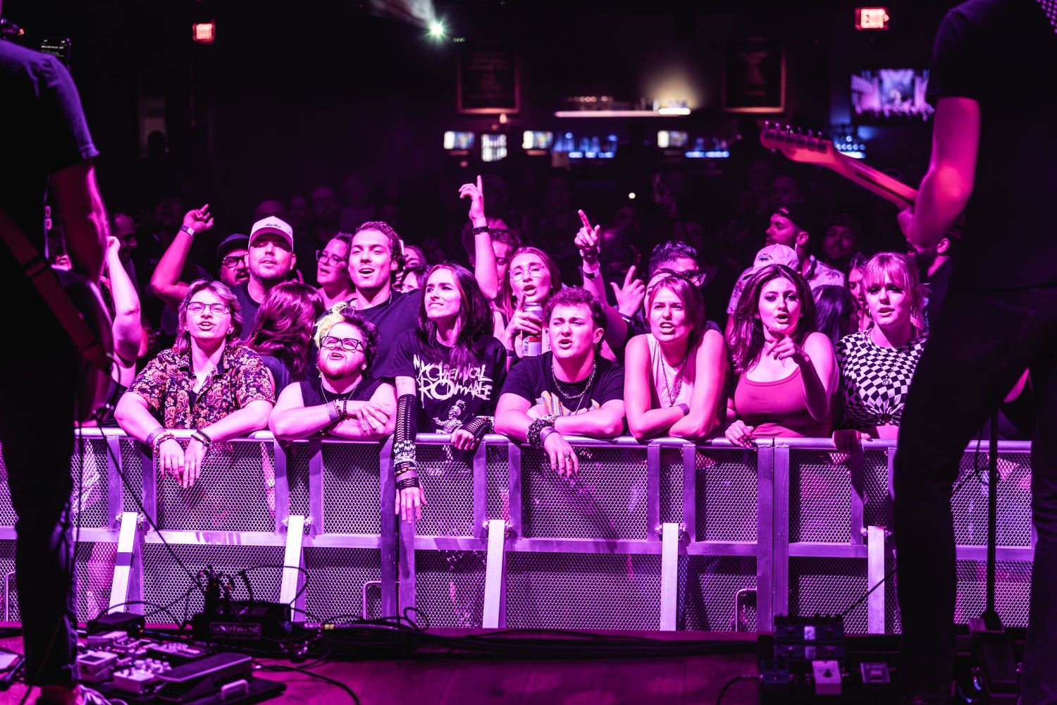 Excited fans at concert barrier reach toward stage under purple lighting.