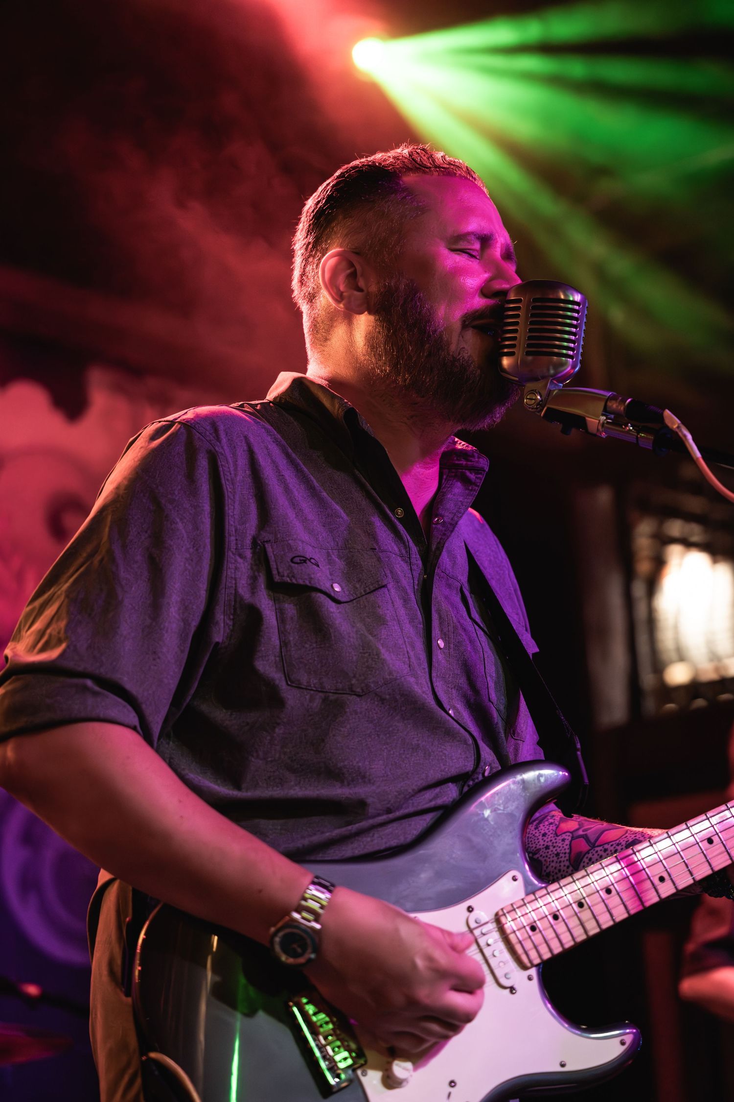 Guitarist sings into retro microphone under dramatic red and green stage lighting.