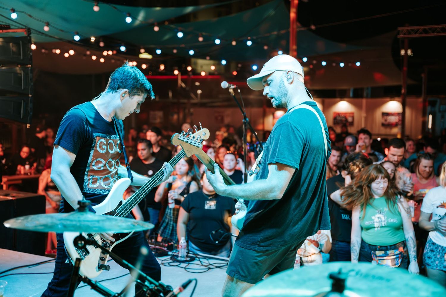 Musicians perform on stage at an intimate venue while an energetic crowd watches under colored lighting.