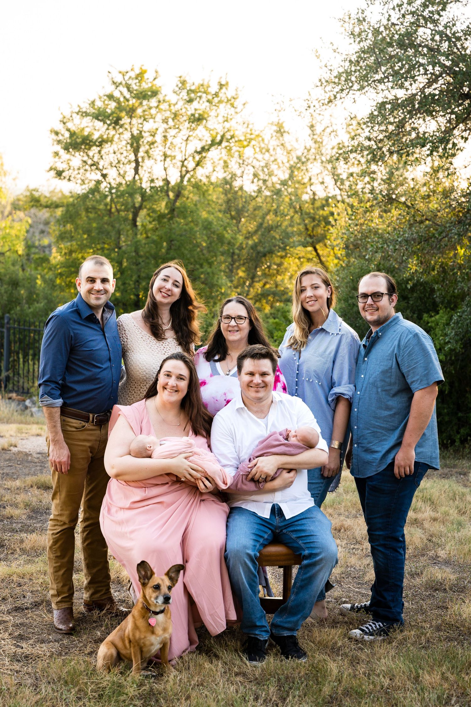 Extended family poses outdoors with a small dog in golden sunset lighting.