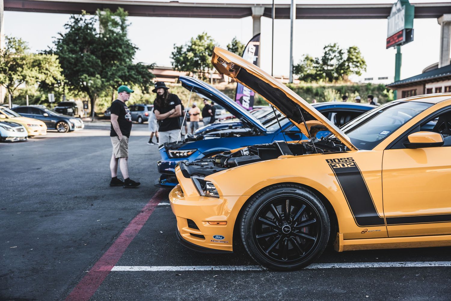 Yellow Ford Mustang with open hood at car show with spectators viewing.