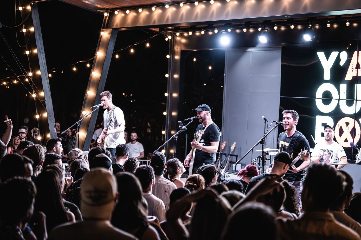 Band performs on illuminated stage with string lights above enthusiastic crowd.
