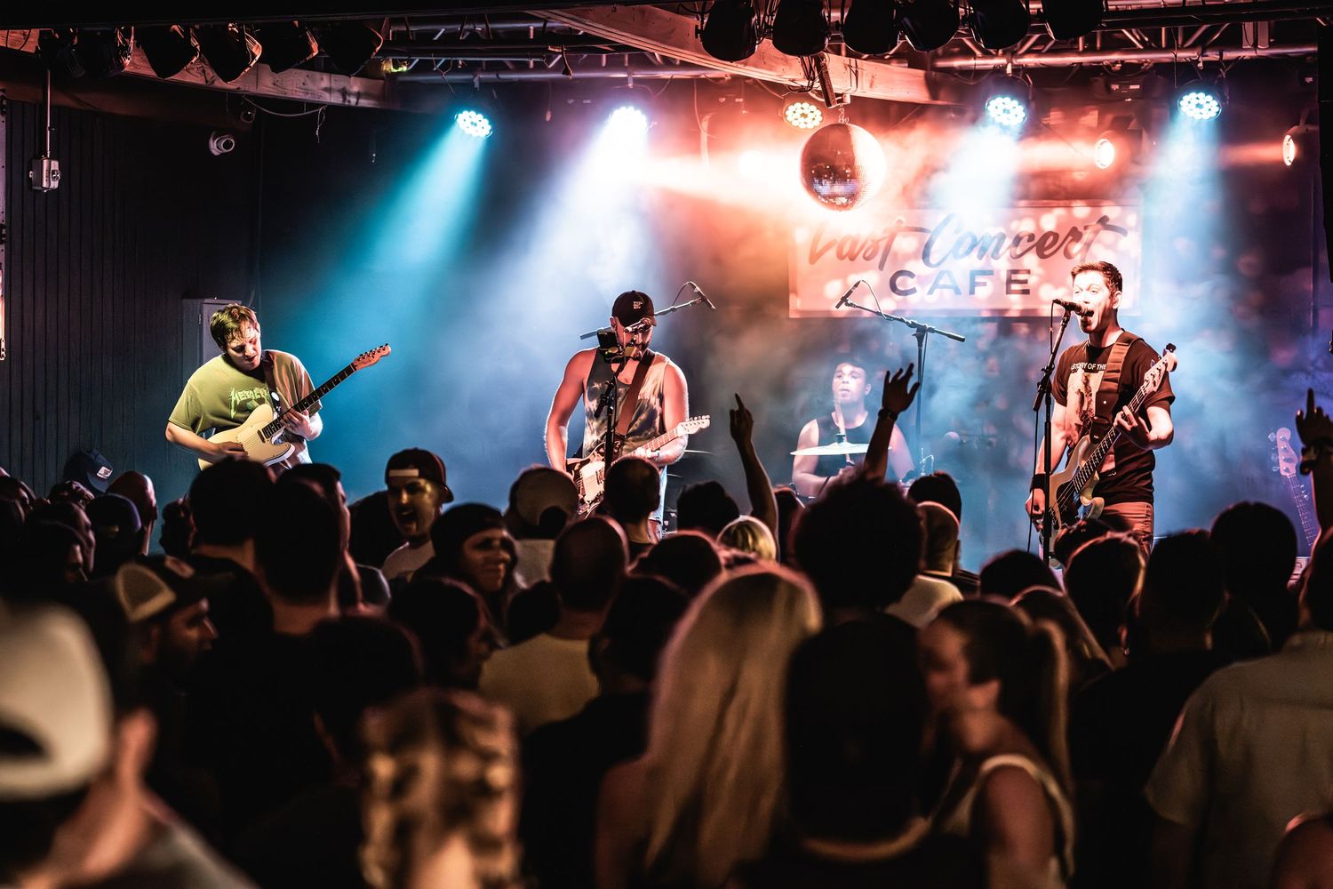 Band performs on stage with colorful blue and orange lighting while crowd watches at intimate concert venue.