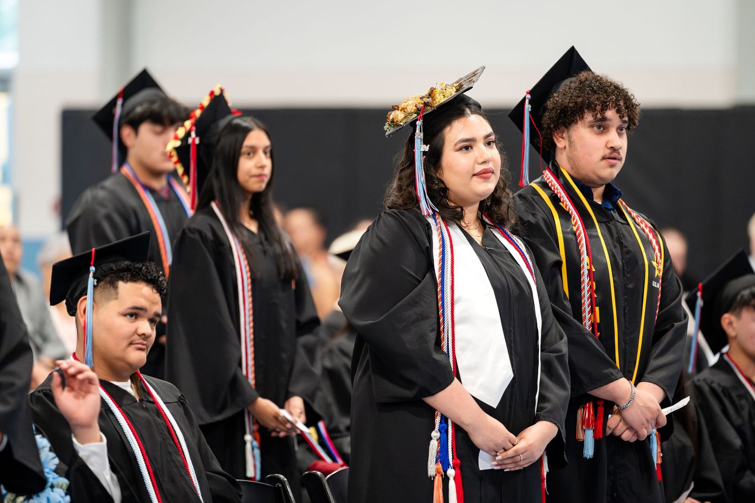 Group of graduates in black caps and gowns stand together during graduation ceremony.