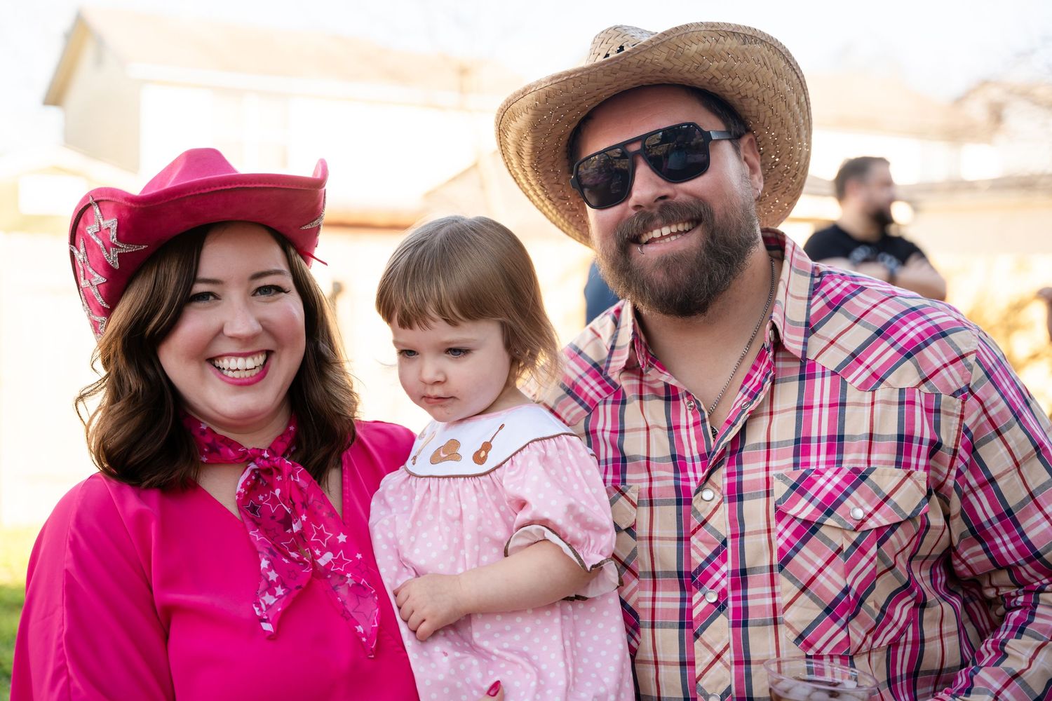 Three people in western attire including a pink cowgirl outfit, straw hat, and plaid shirt pose together outdoors.