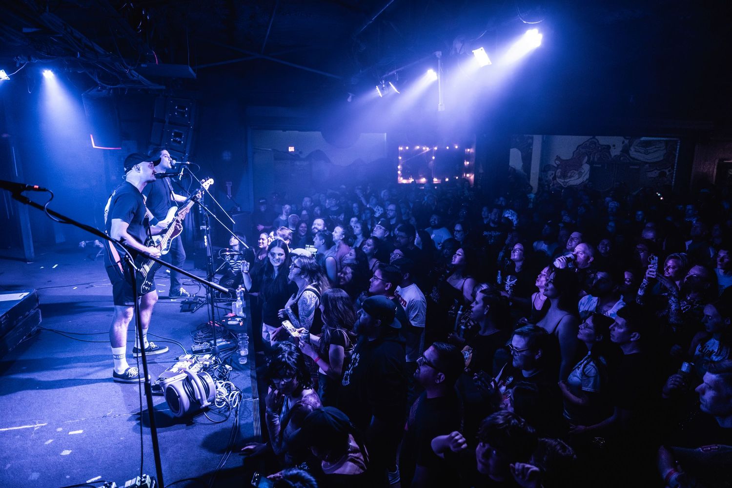Musicians perform on stage at a crowded concert venue with dramatic blue lighting and enthusiastic audience members below.