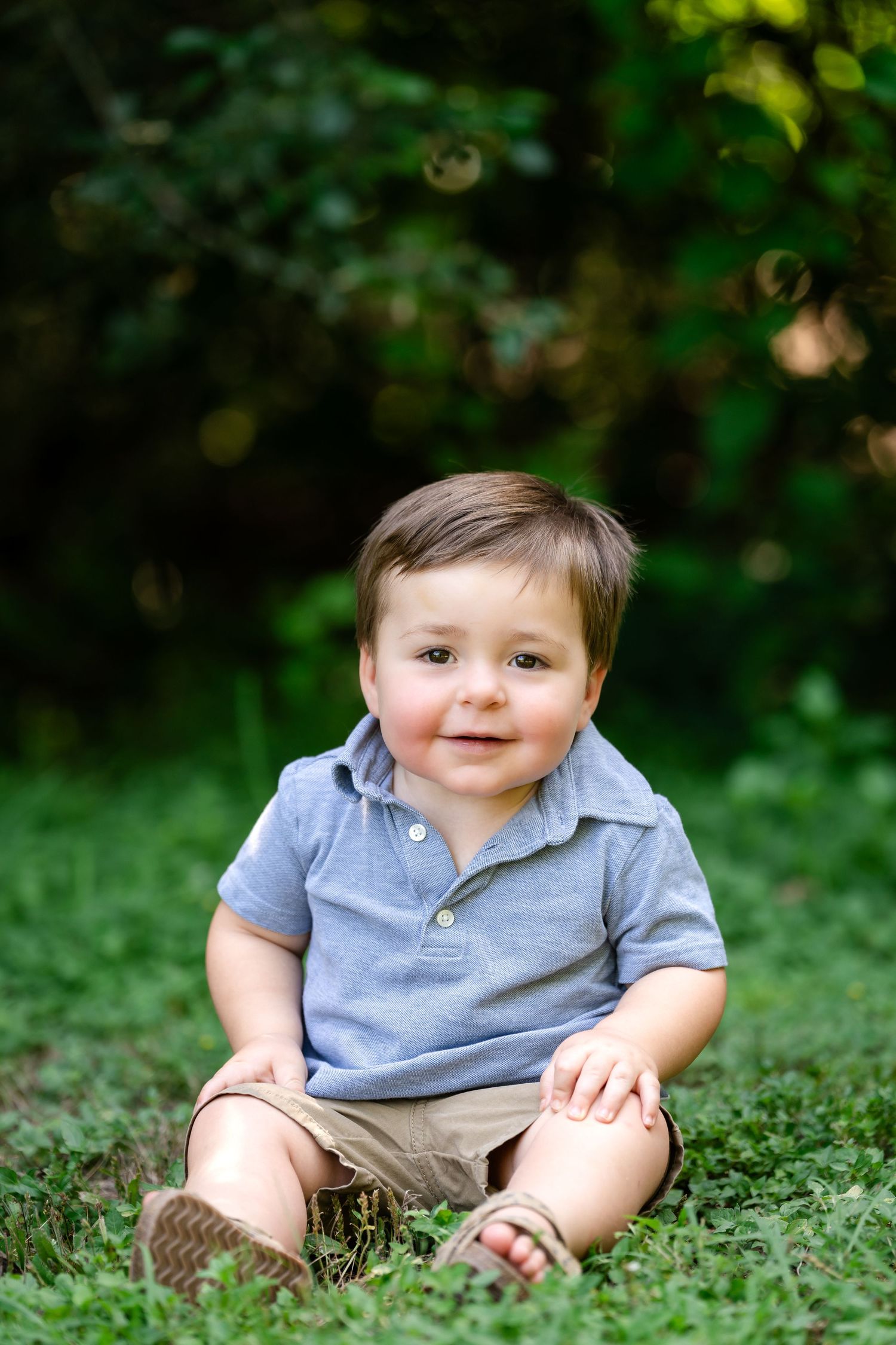 A smiling toddler in blue polo sitting on grass with bokeh lighting effects.