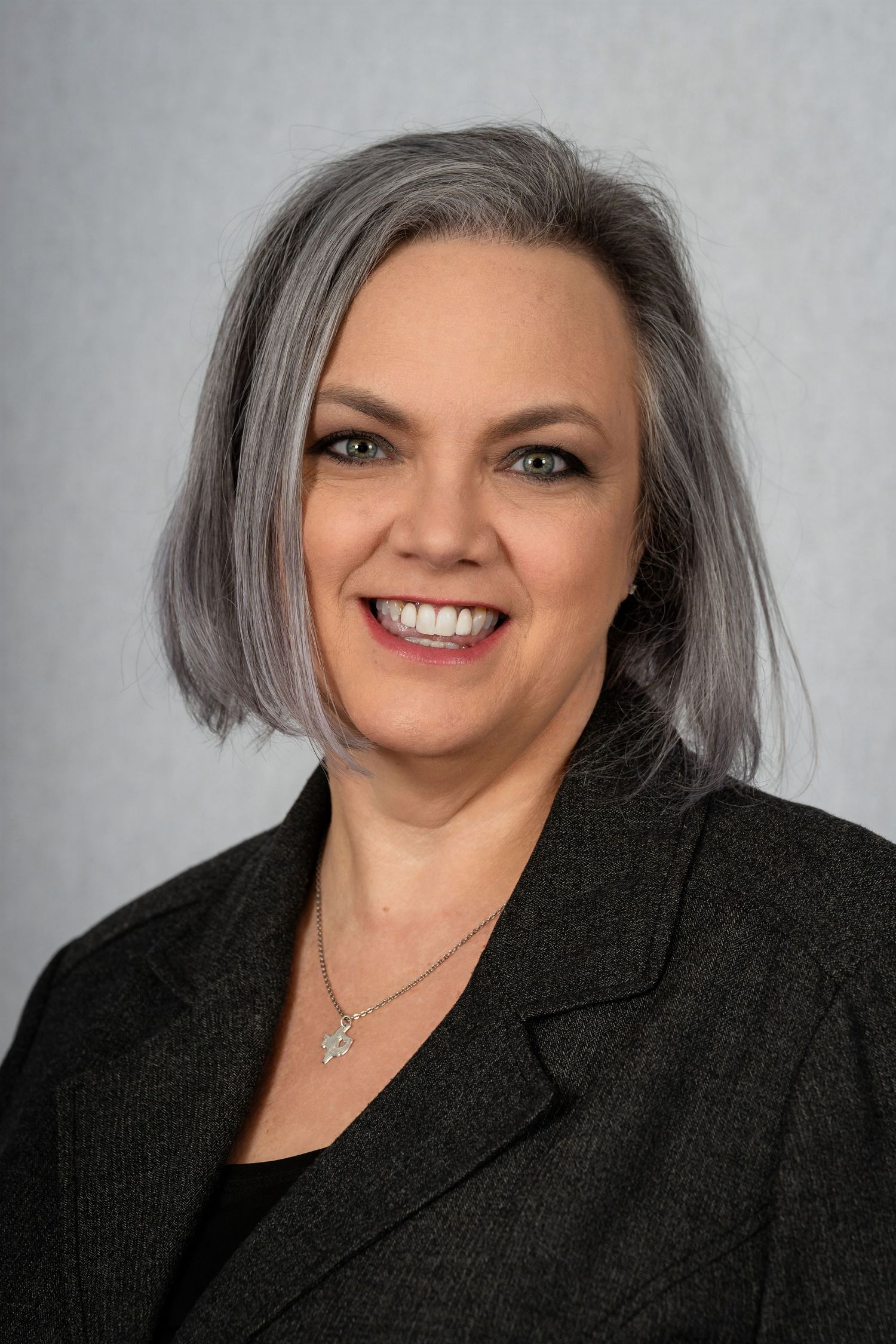 Professional headshot of a smiling woman with silver hair wearing a black blazer against a gray background.
