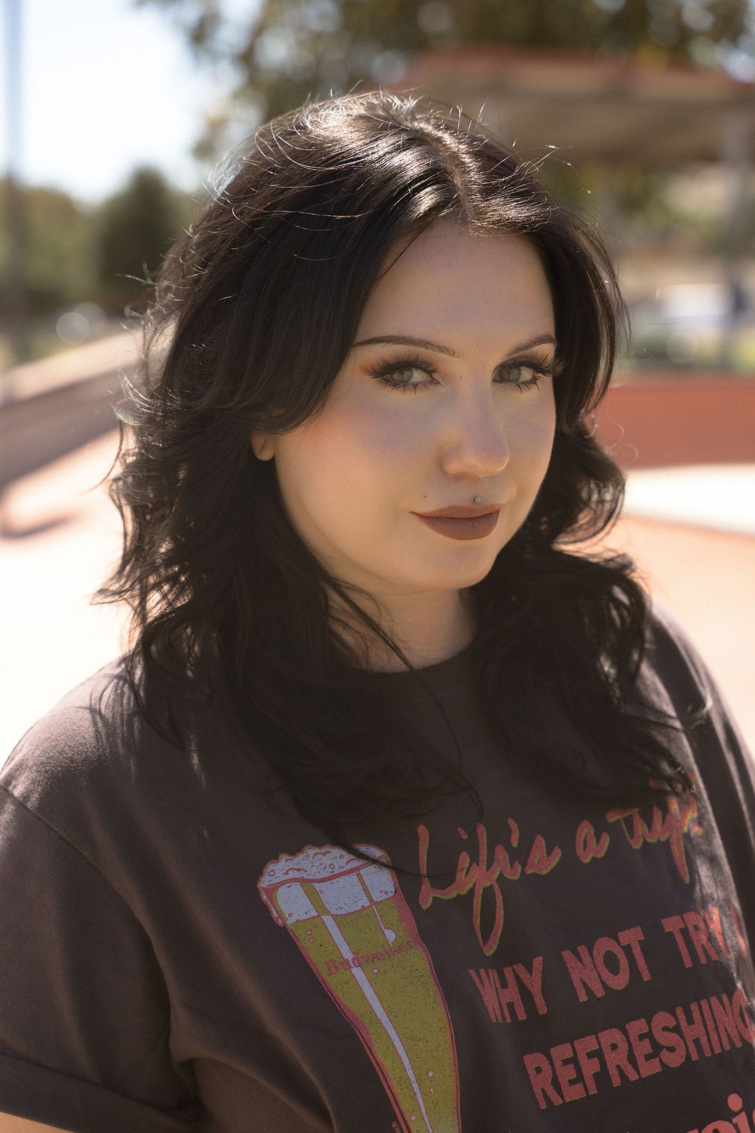 A person wearing a dark graphic t-shirt with text about refreshments poses outdoors.