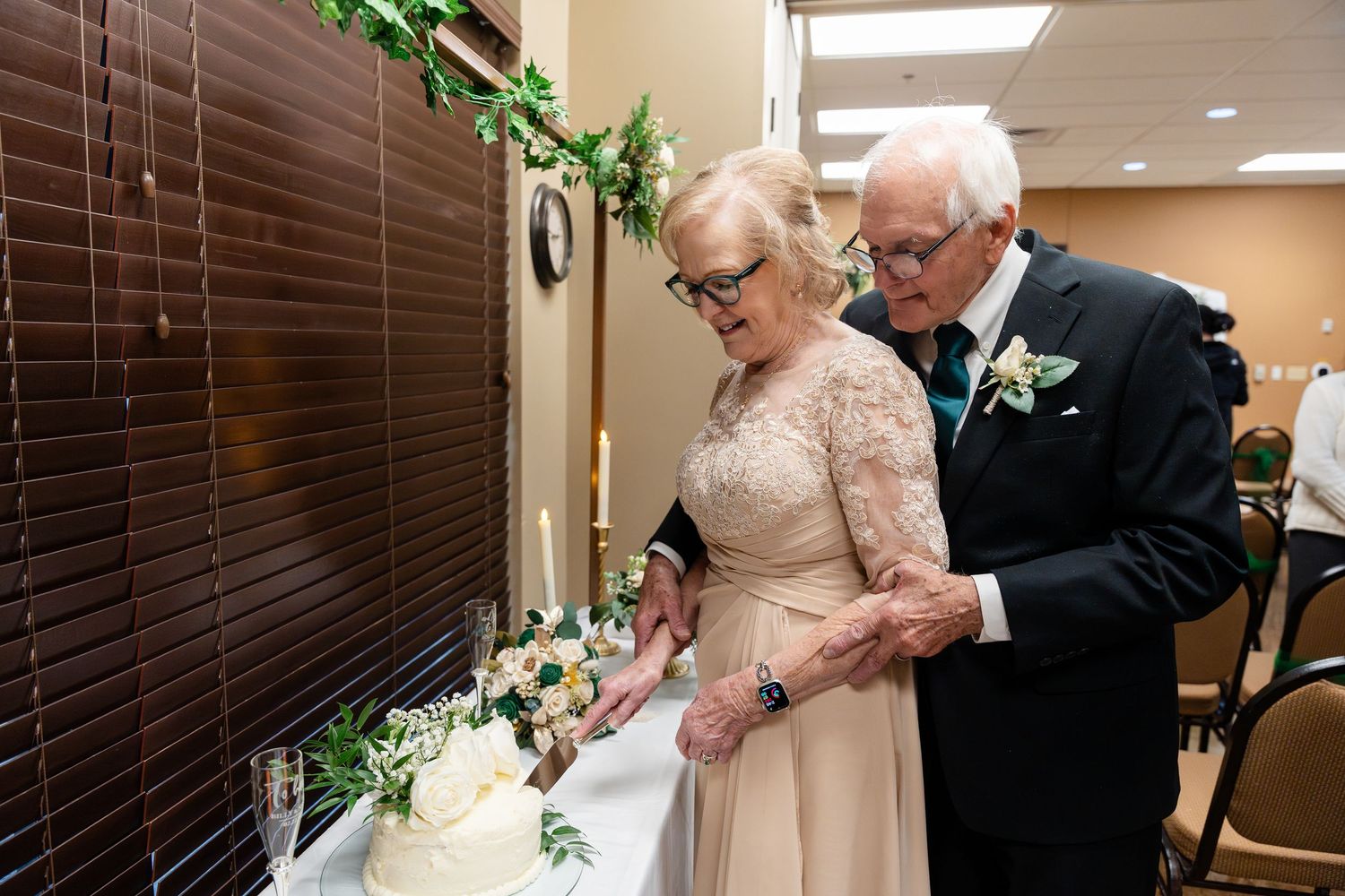 An elderly couple in formal attire cuts their wedding cake together in a reception hall.