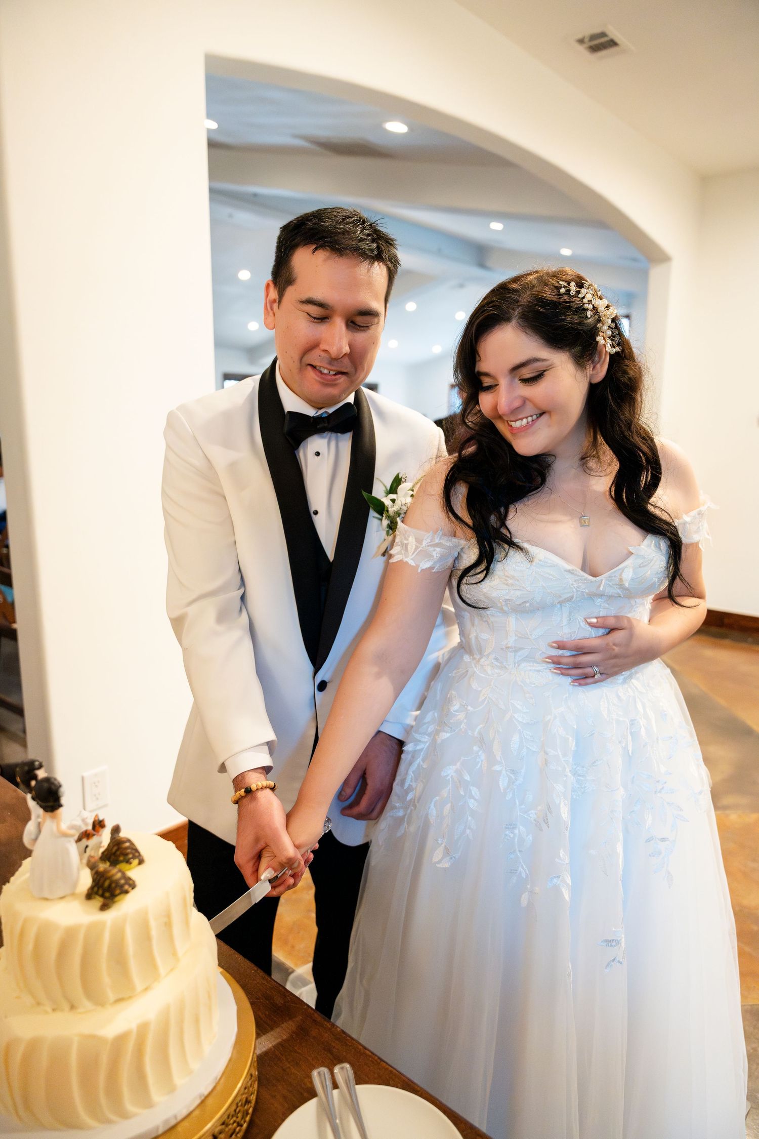 A romantic moment between newlyweds as they cut their elegant white wedding cake together in a bright indoor venue.
