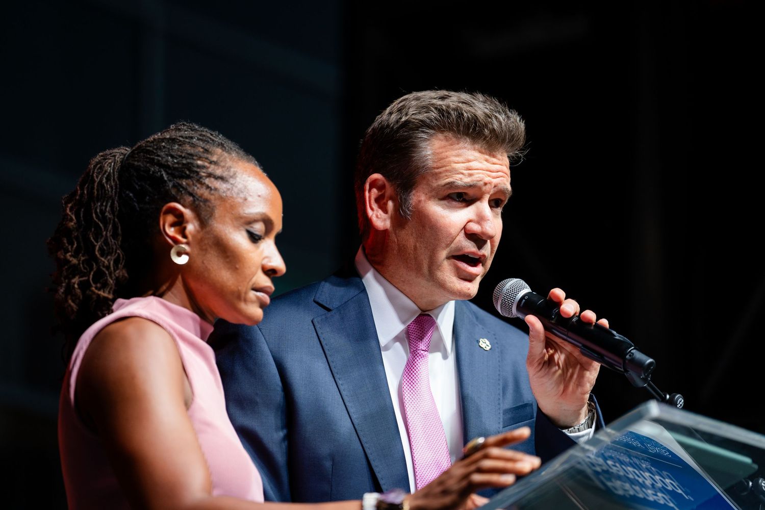 Two speakers addressing an audience from a podium with dramatic stage lighting.