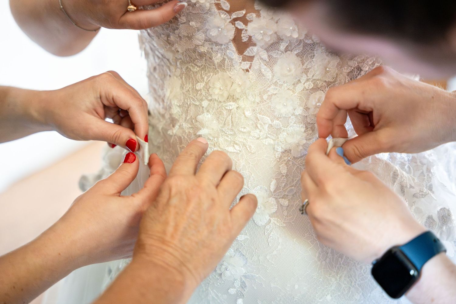 Multiple hands work together to adjust intricate details on a white lace wedding dress.
