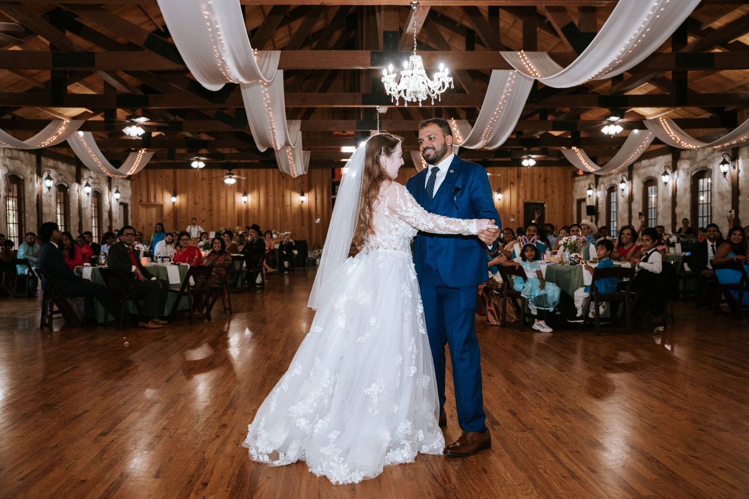 A couple shares their first dance in an elegant wood-beamed reception hall with draped fabric decor.