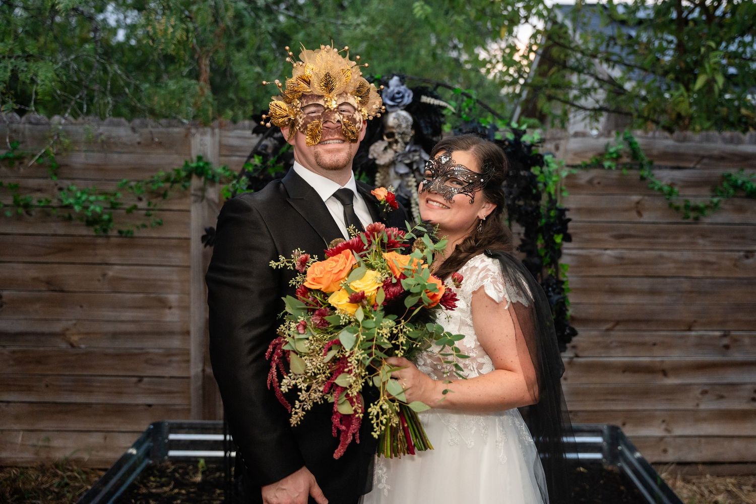 Wedding couple poses with autumn-colored bouquet wearing decorative masks against wooden backdrop.