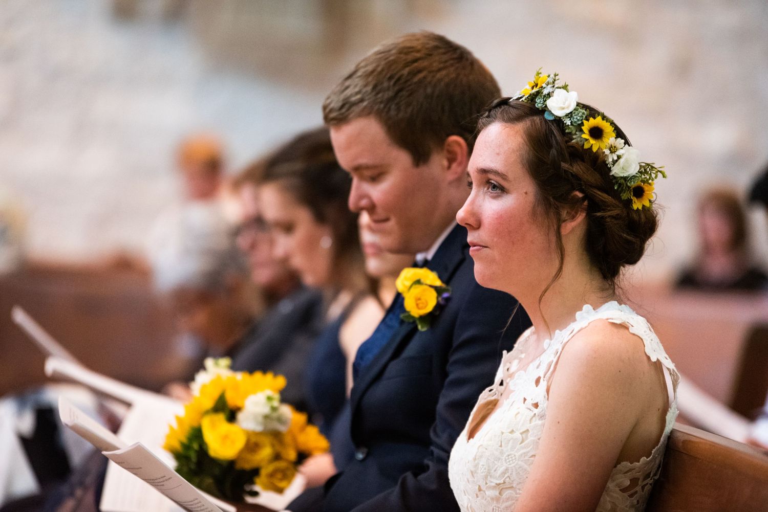 A couple sits in a church pew during a wedding ceremony with yellow floral bouquets and decor.