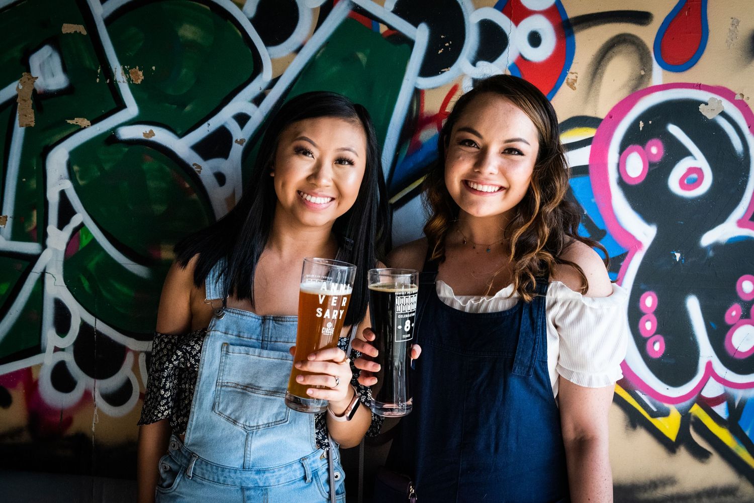 Two friends enjoying craft beers together in front of colorful graffiti wall art.